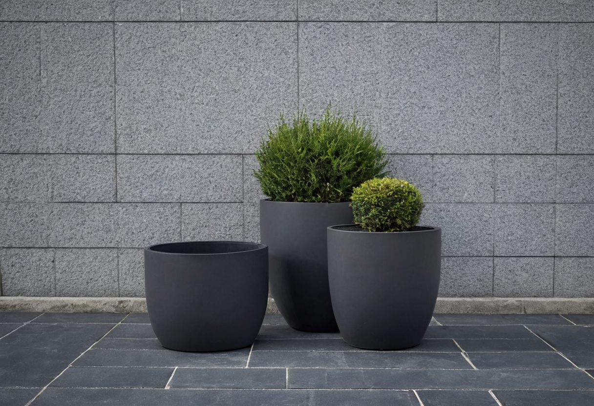 Three dark round planters of differing heights and widths on dark paving against a gray wall; the left is empty and the others hold shrubs.