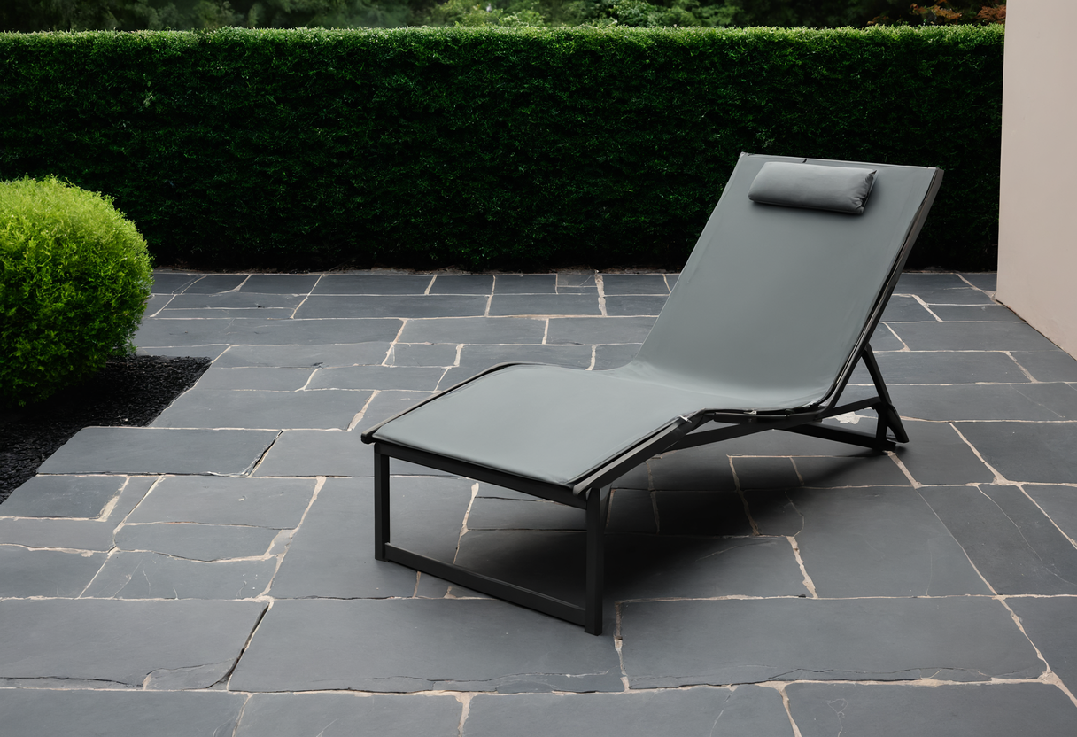 Single grey lounge chair on flagstone paving with open ground around it and a hedge behind
