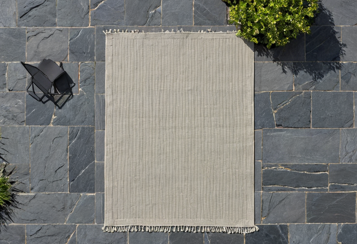 Overhead view of a pale rectangular woven rug on dark stone paving with an empty black chair to the side.